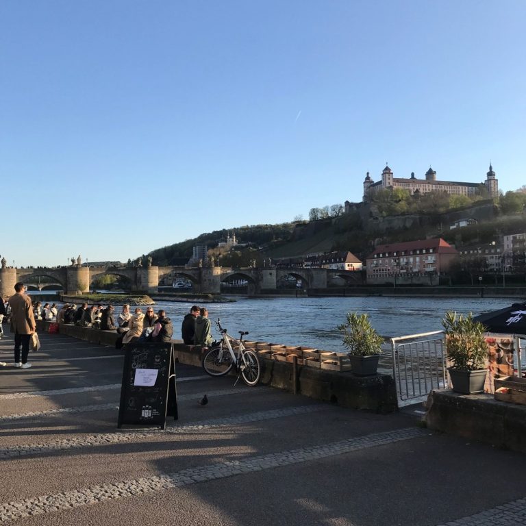 Menschen sitzen am Main bei sommerlichem Wetter. Im Hintergrund die Festung und die Alte Mainbrücke.
