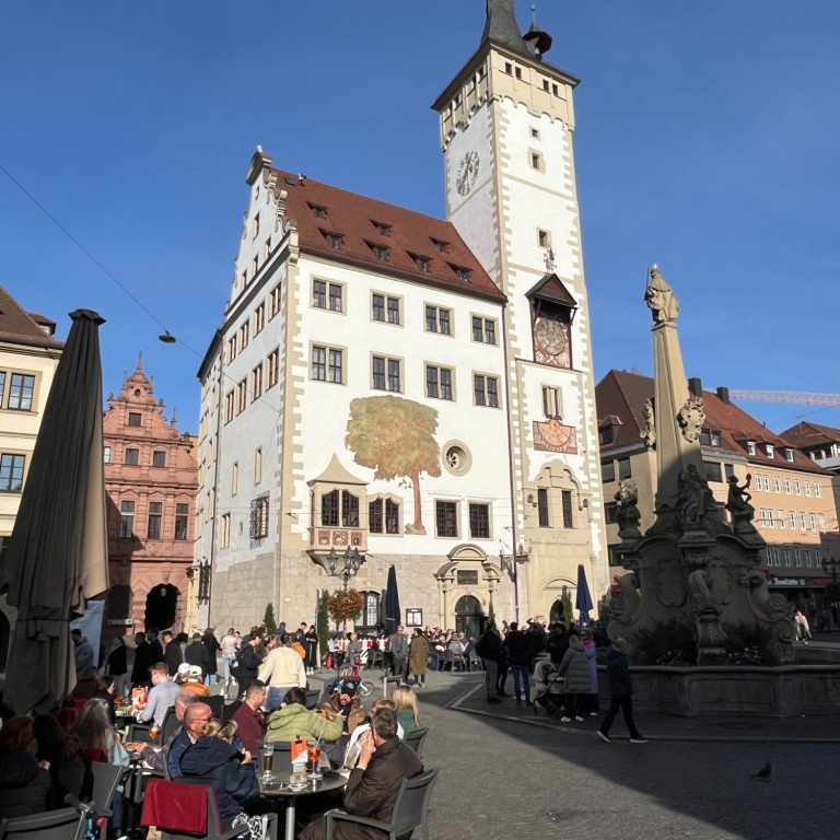 Würzburger Rathaus (genannt Grafeneckart)  im Sommer. Im Vordergrund Menschen im Straßencafe und ein Brunnen.